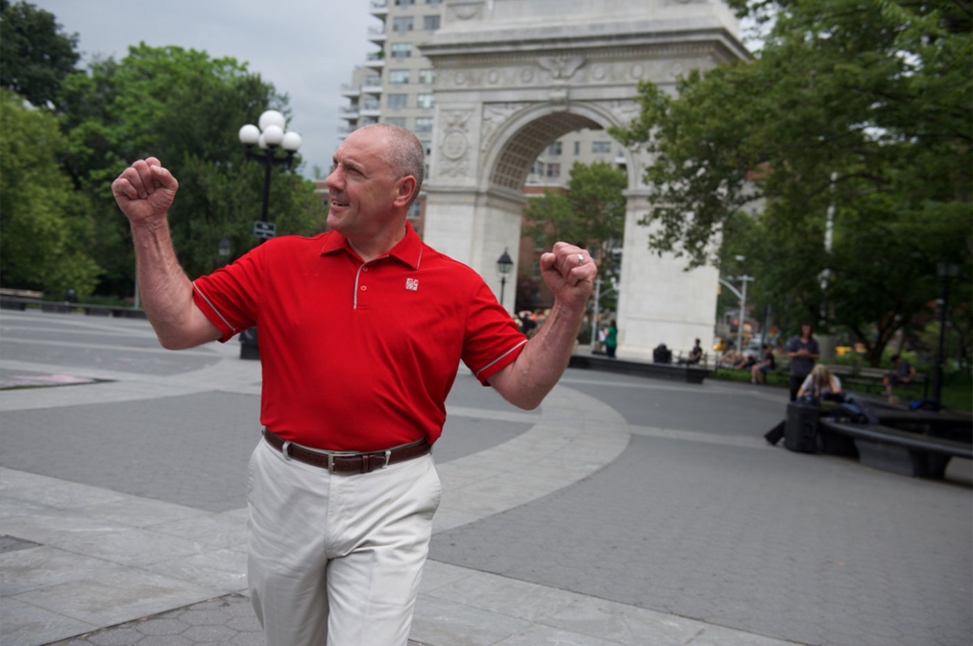 A man in a red Chick-fil-A polo raises his arms in celebration while walking through a city plaza with a large stone arch in the background.