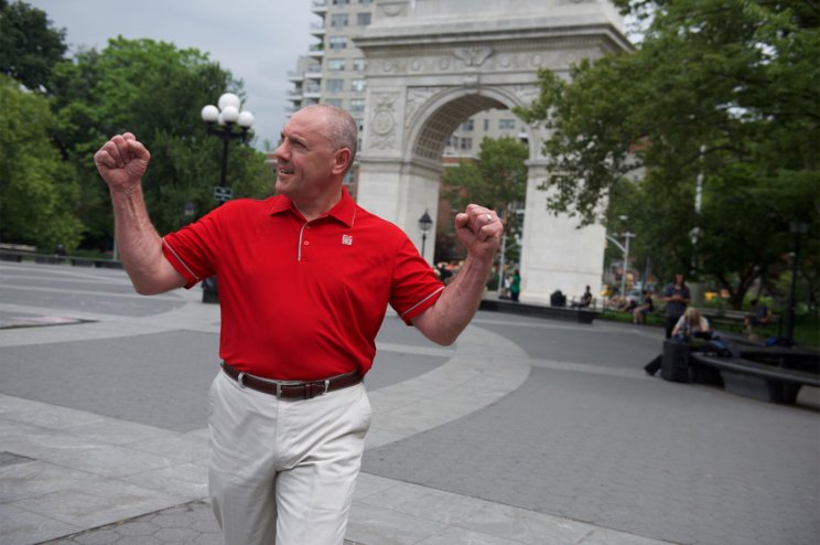 A man in a red Chick-fil-A polo raises his arms in celebration while walking through a city plaza with a large stone arch in the background.