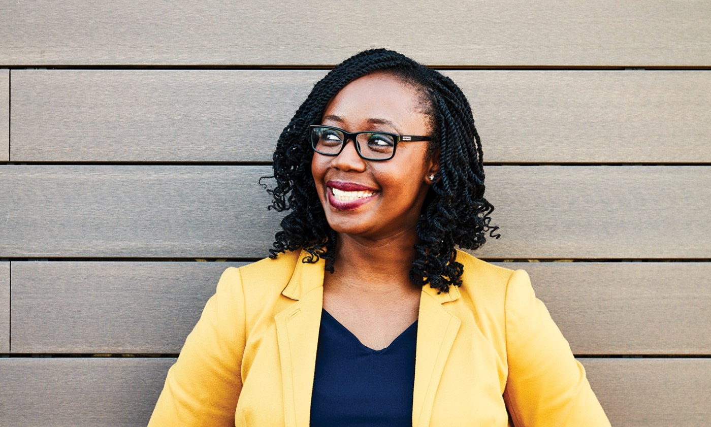 Woman in a yellow blazer smiling in front of wooden panels.