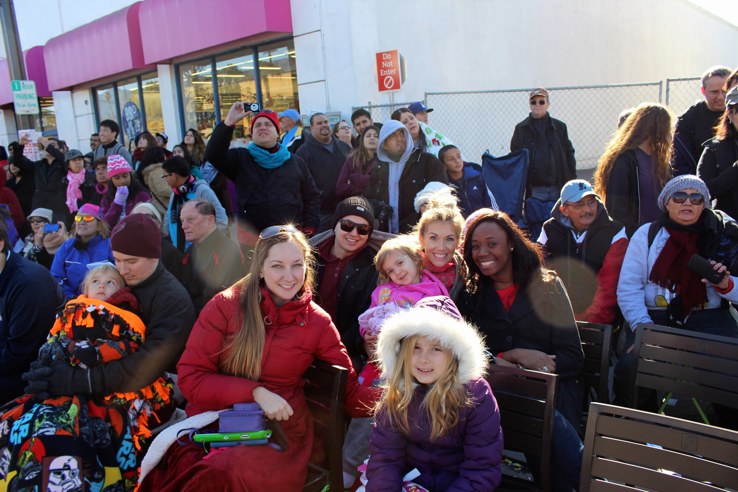 A cheerful crowd dressed in winter clothing gathers outdoors, with families and children seated and smiling during a public event.