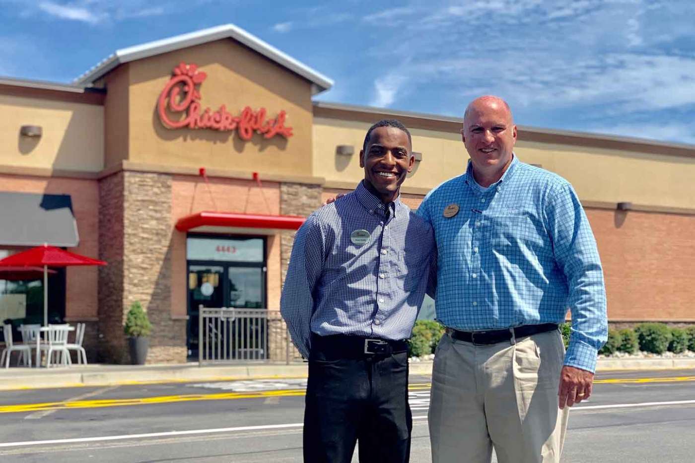 Two Chick-fil-A team members stand smiling in front of a Chick-fil-A restaurant on a sunny day.
