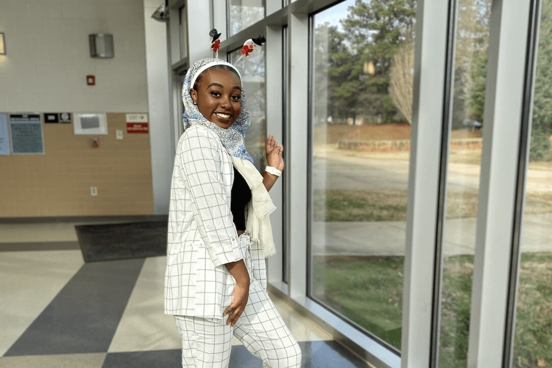Adar Hussein smiles while posing indoors in a white plaid suit and blue-patterned headscarf, standing near large windows with natural light.
