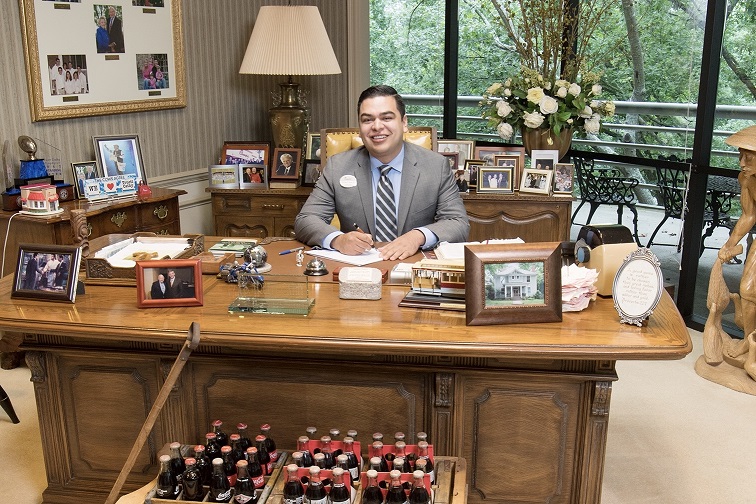 Enrique Baca, wearing a suit and Chick-fil-A name tag, smiles while seated at a desk surrounded by family photos and memorabilia in an office with large windows.