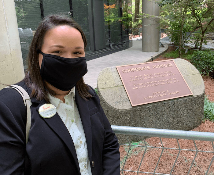 Woman in a mask standing by a stone plaque outside a modern building.