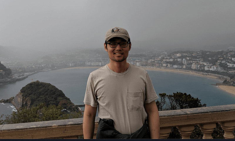A person stands smiling in front of a coastal bay with a cityscape and hills in the background.