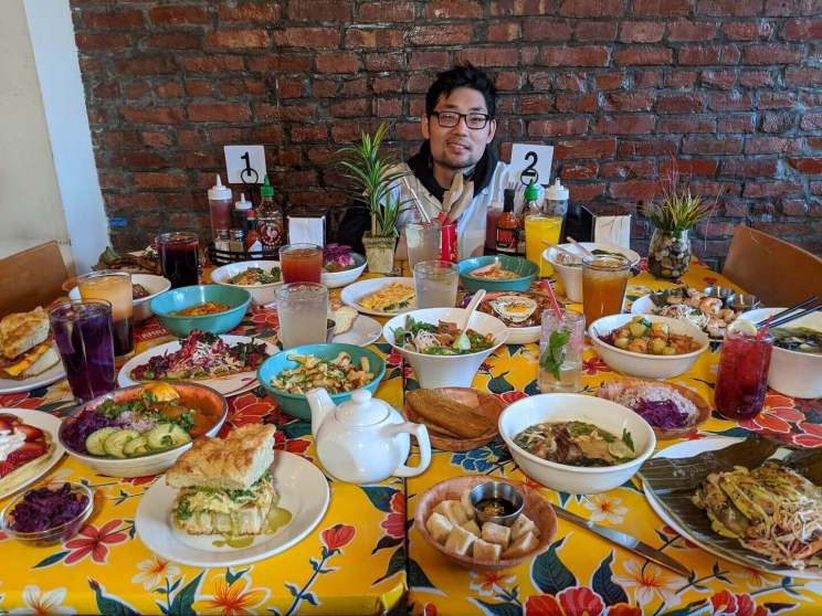 A colorful table filled with diverse dishes and beverages, with a man seated in the center against a brick wall.