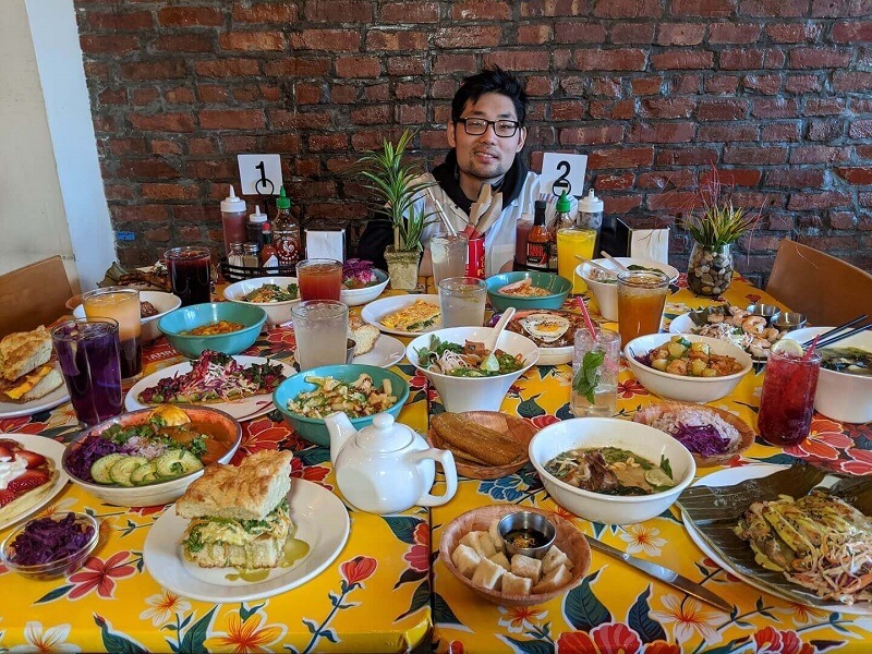 A colorful table filled with diverse dishes and beverages, with a man seated in the center against a brick wall.