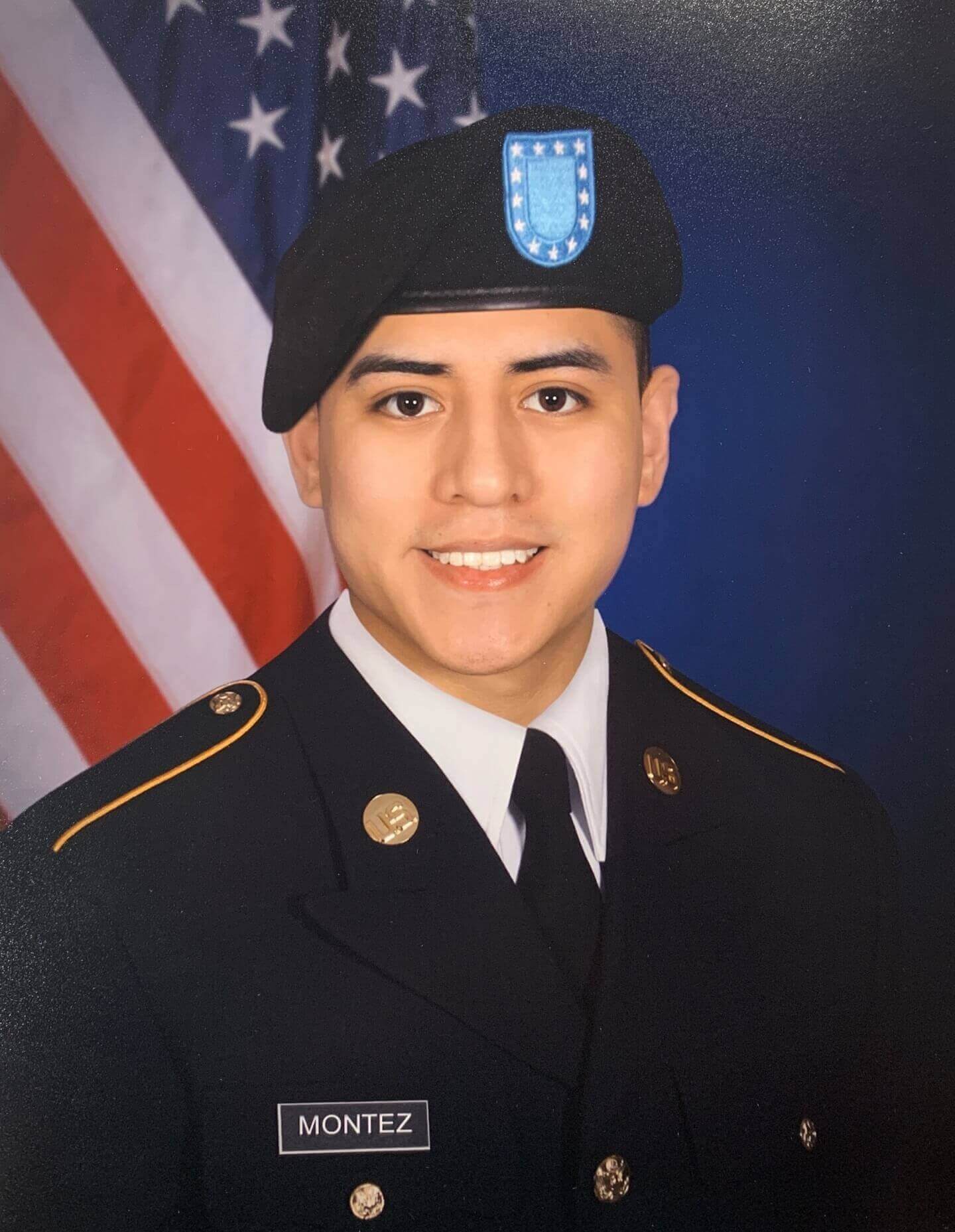 A young man in a formal U.S. Army uniform smiles in front of an American flag backdrop. His name tag reads “Montez.”