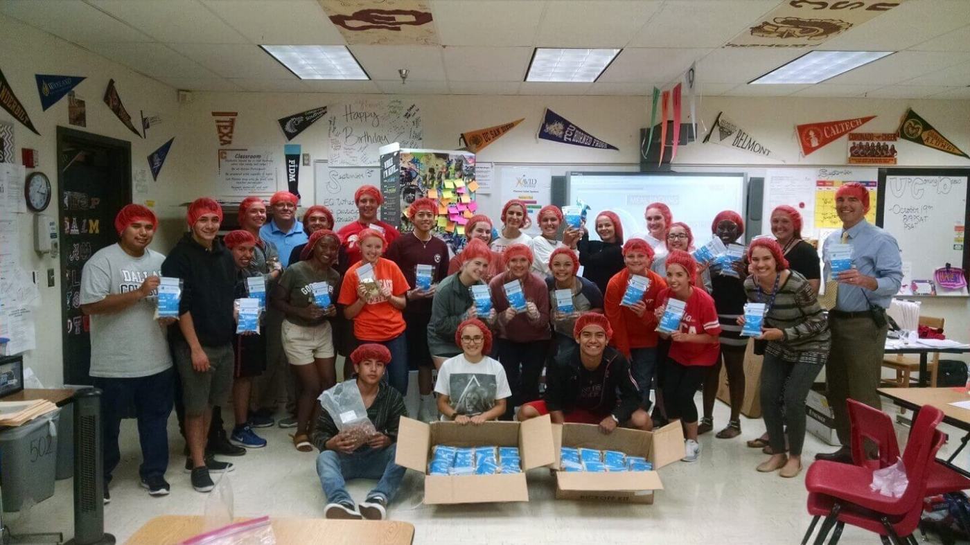 A large group of students and adults wearing red hairnets pose in a classroom holding meal packages they assembled for a service project.