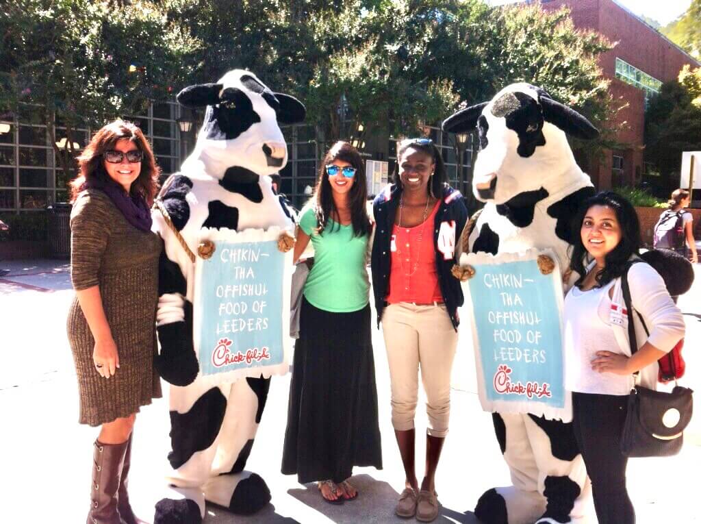 Four women pose outdoors with two people in cow costumes holding signs that say “Chikin—the Offishul Food of Leeders,” promoting Chick-fil-A.