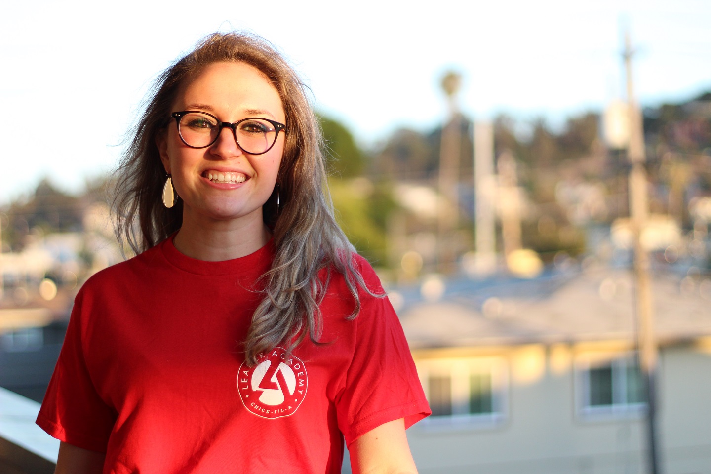 A smiling young woman wearing a red “Leader Academy Chick-fil-A” T-shirt stands outside with houses and power lines in the background.