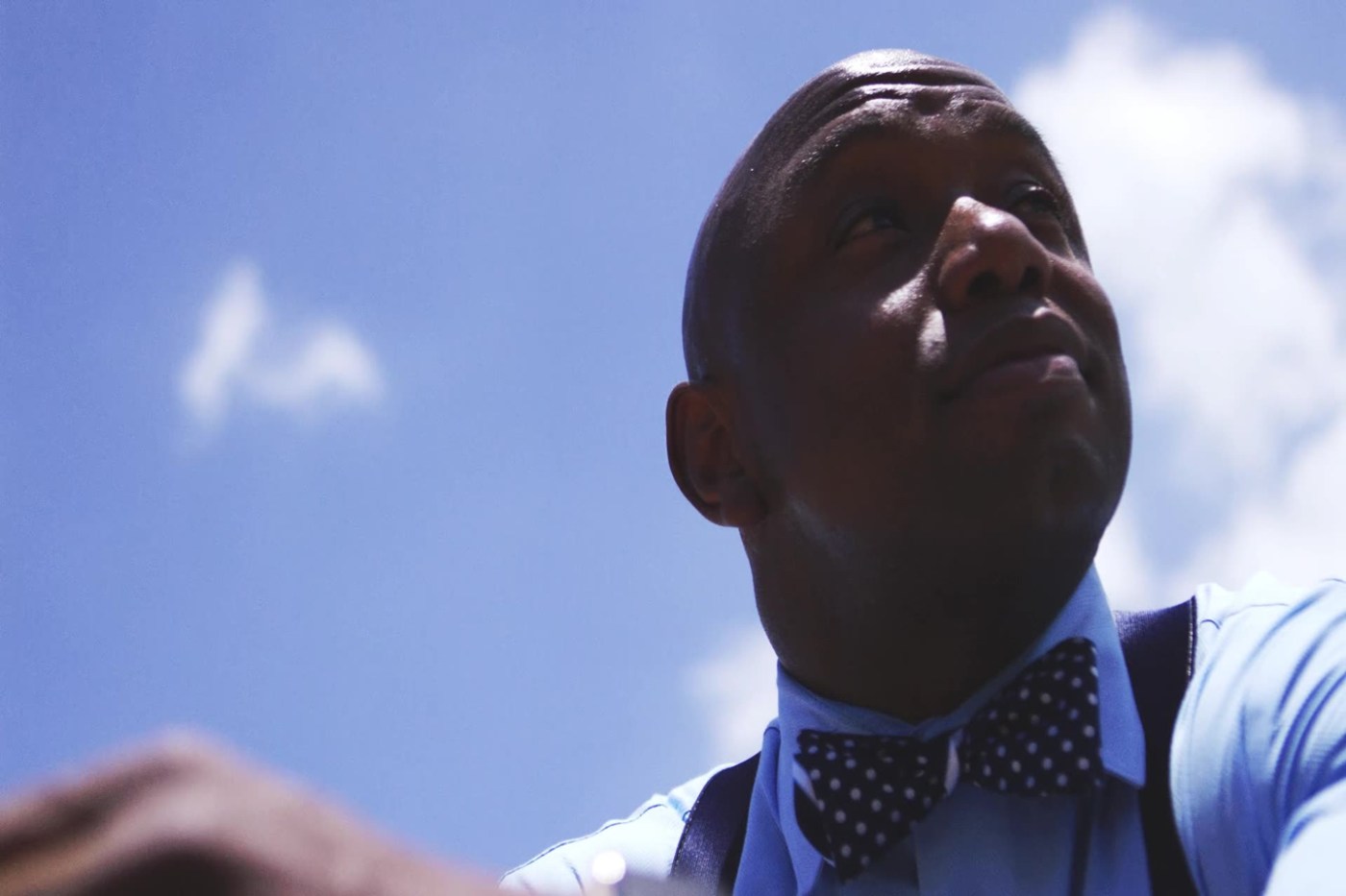 Person in a blue shirt and polka dot bow tie, looking upward against a blue sky with clouds.