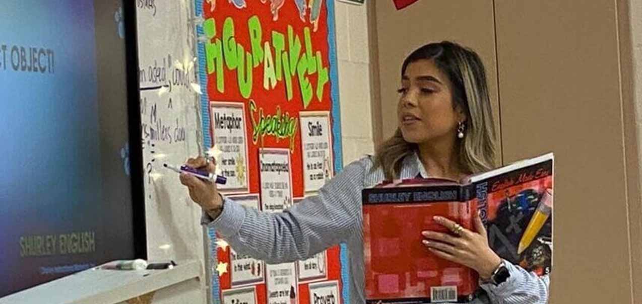 A teacher writes on a whiteboard while holding an English textbook in a classroom decorated with colorful posters.