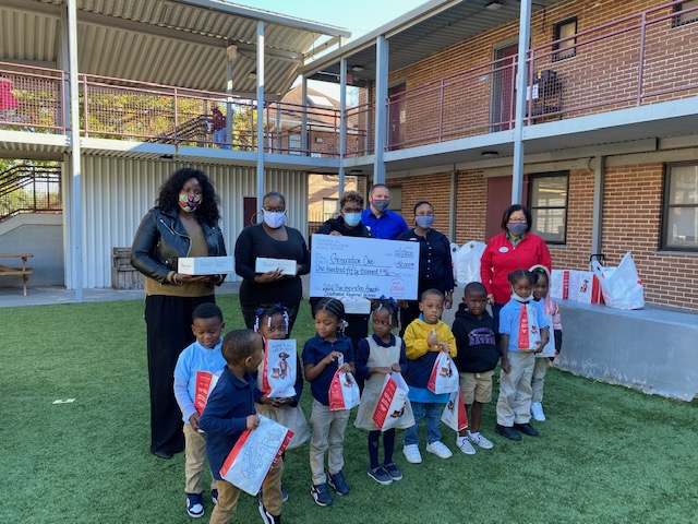 A group of adults and children pose with a large check and gift bags in a courtyard.