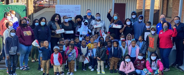 A group of adults and children pose with a large check and gift bags in a courtyard.