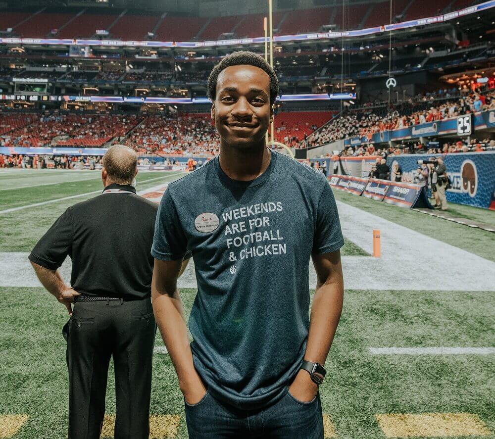 Smiling Chick-fil-A team member stands on a football field wearing a shirt that reads “Weekends are for football & chicken.”