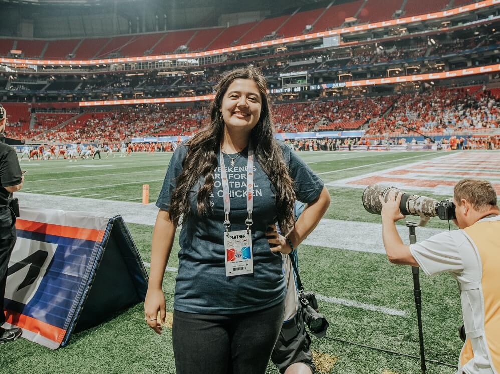 Smiling Chick-fil-A team member stands near the end zone of a football field wearing a shirt that reads “Weekends are for football & chicken.”