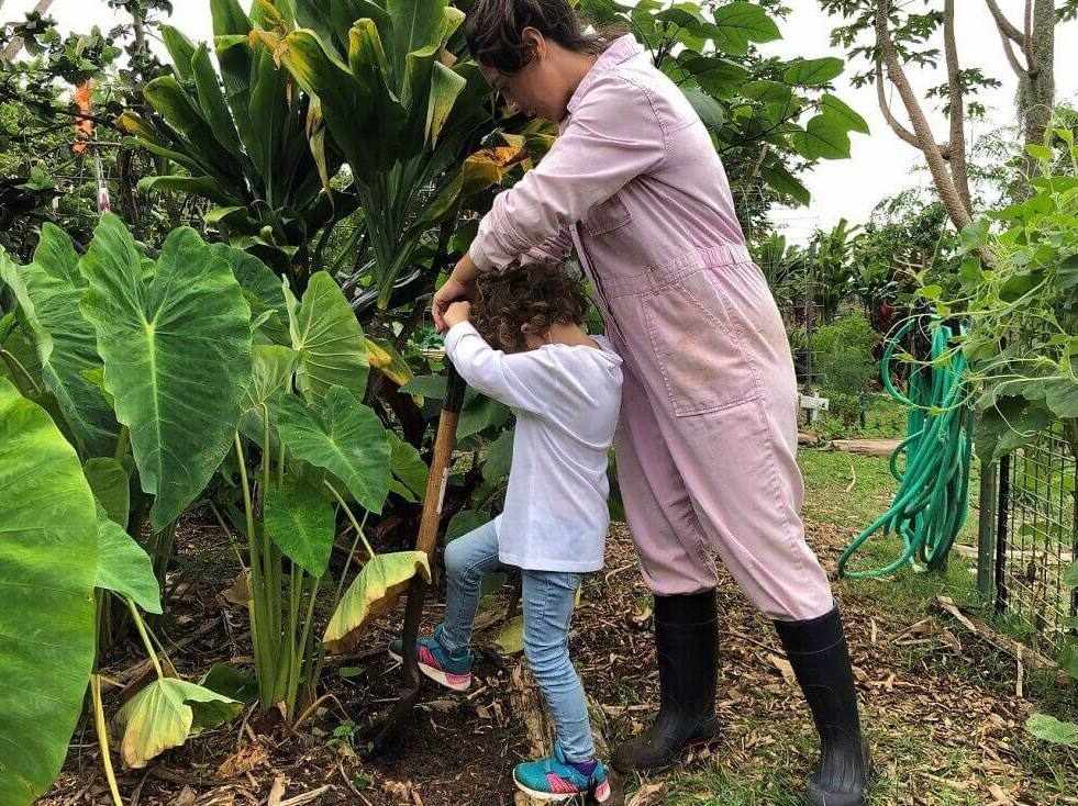 A woman and a young girl work together to dig in a lush garden surrounded by large green plants.