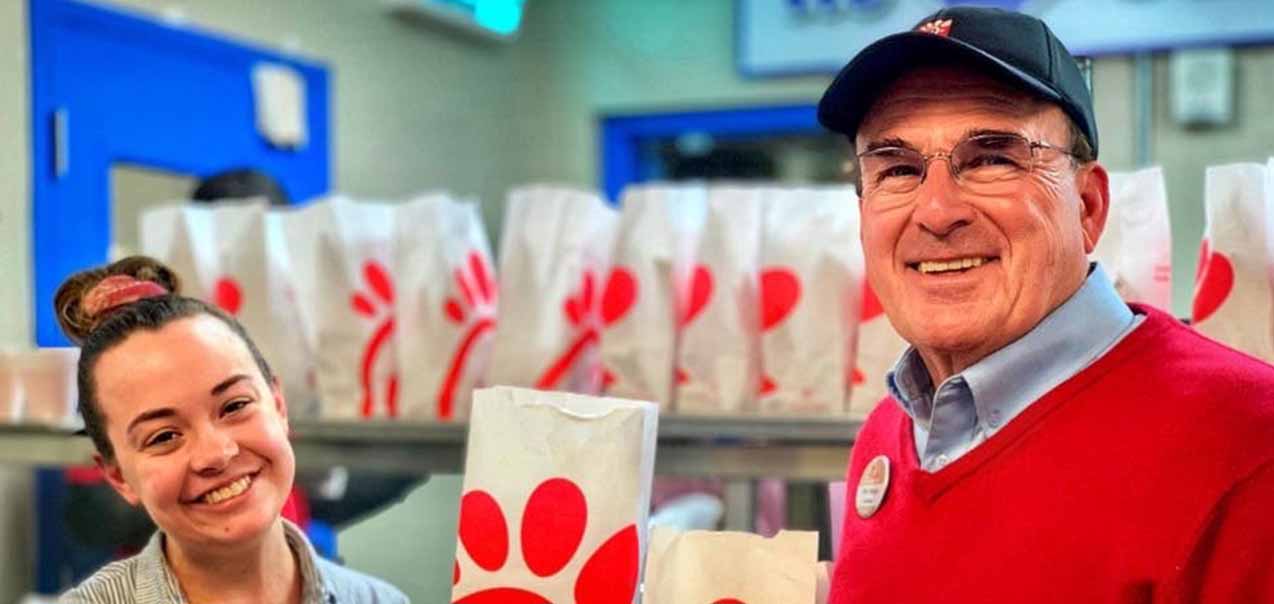 Two smiling Chick-fil-A team members standing in front of a counter lined with Chick-fil-A branded takeout bags.