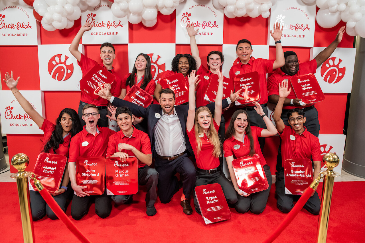 A group of Chick-fil-A scholarship recipients in red uniforms pose joyfully on a red carpet with award plaques in front of a red and white Chick-fil-A backdrop decorated with balloons.