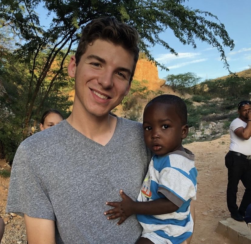 A young man in a gray t-shirt holds a small child in a striped shirt outdoors, with trees and rocky terrain in the background.