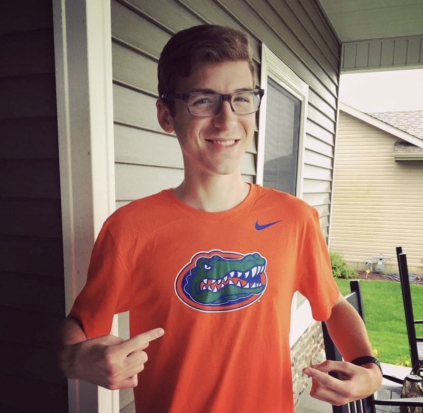 A smiling young man wearing glasses and an orange University of Florida Gators shirt stands on a porch, pointing at the logo.