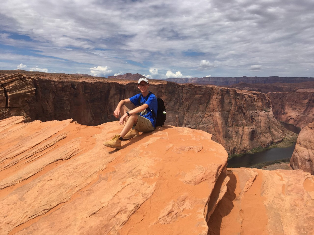 A young man in a blue shirt and tan boots sits on the edge of a red rock cliff with a canyon and river in the background under a cloudy sky.