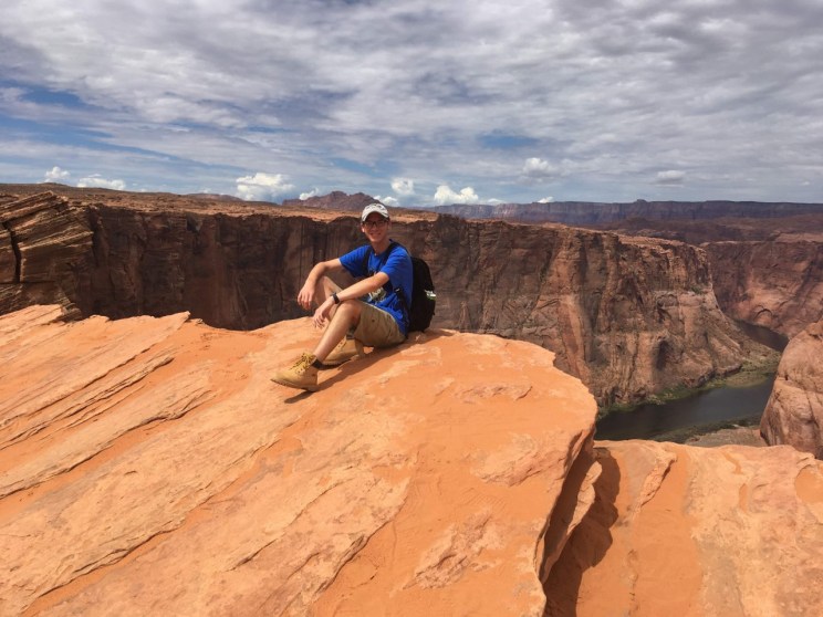 A young man in a blue shirt and tan boots sits on the edge of a red rock cliff with a canyon and river in the background under a cloudy sky.