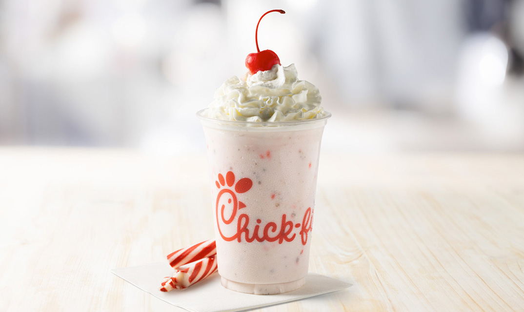 Chick-fil-A peppermint milkshake in a clear cup, topped with whipped cream and a cherry, with peppermint sticks placed beside the cup.