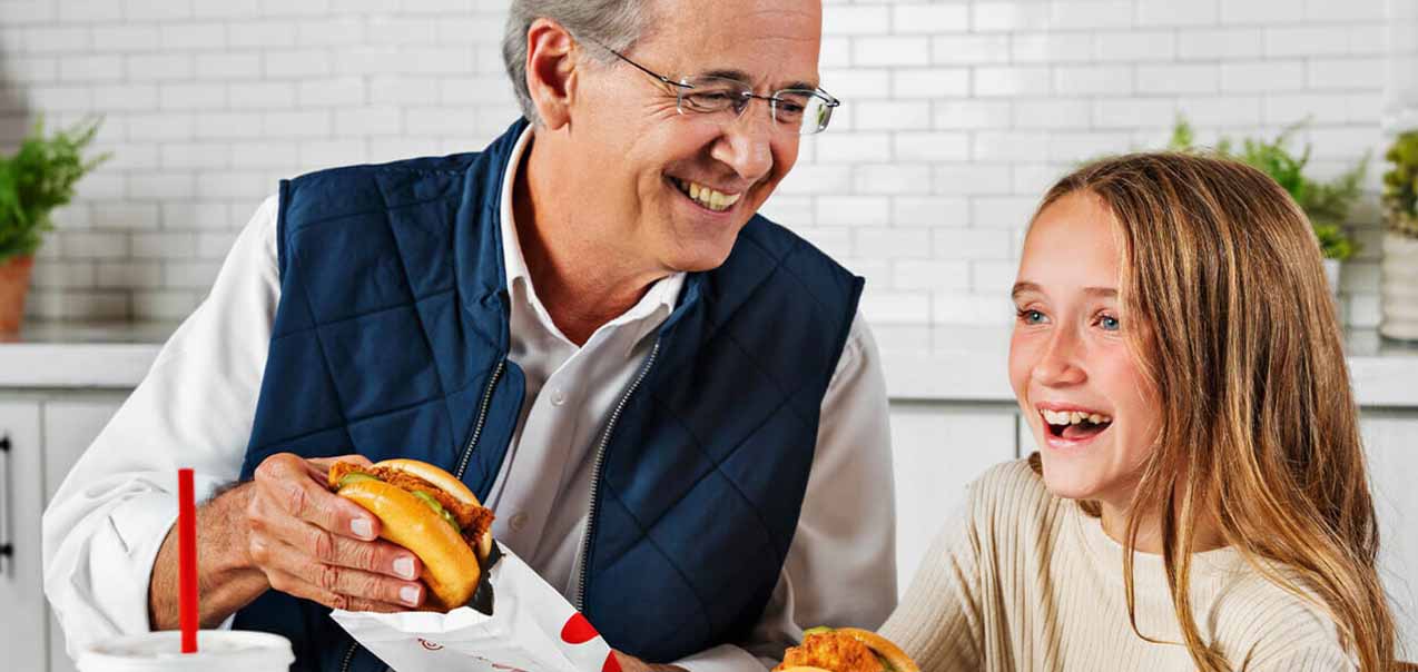 A man and a girl sitting next to each other eating the Chick-fil-A® Chicken Sandwich and smiling
