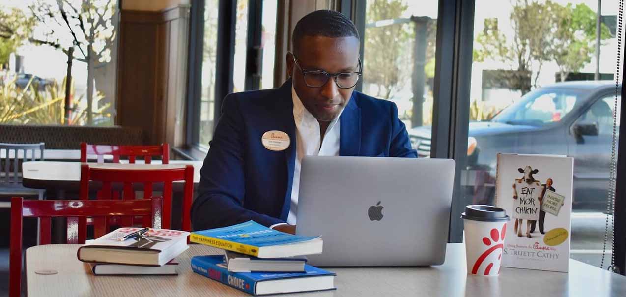 Jon Hooper works on a laptop at a table with books, a Chick-fil-A cup, and an “Eat Mor Chikin” display in a restaurant dining area.