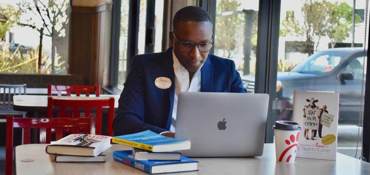 Jon Hooper works on a laptop at a table with books, a Chick-fil-A cup, and an “Eat Mor Chikin” display in a restaurant dining area.