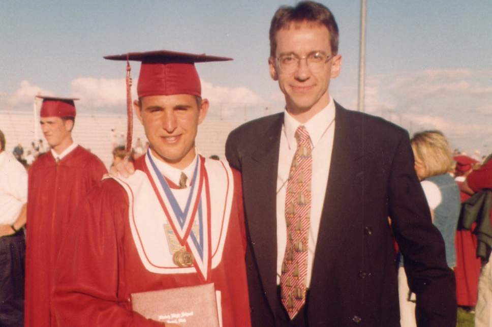 Vince Merrell in a red cap and gown stands next to an adult in a suit, outdoors on a sunny day.

