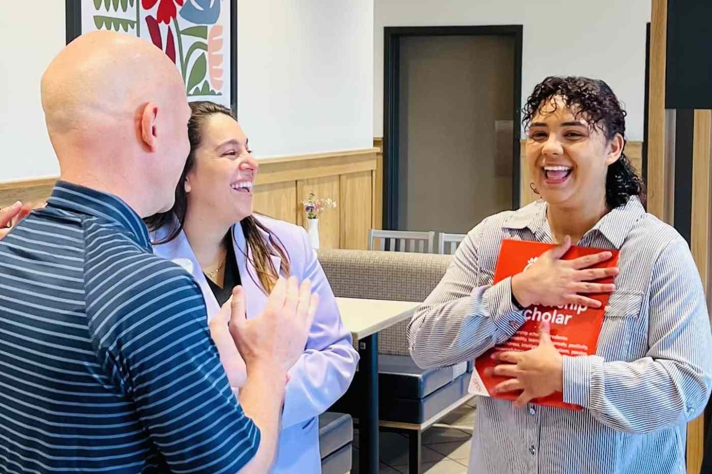 Smiling woman holds a red Chick-fil-A Leadership Scholarship folder to her chest while being congratulated by two others in a restaurant setting.