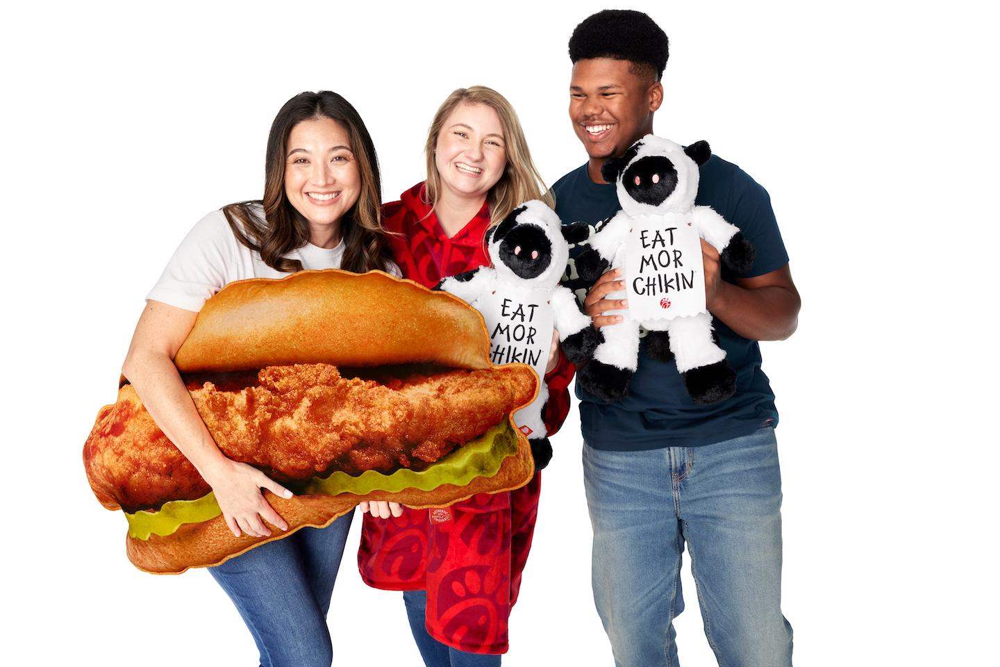 Three people posing with a chicken sandwich cutout and plush cow toys.