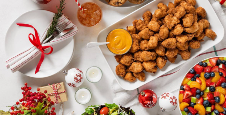 Table with a platter of Chick-fil-A® Nuggets Tray, a bowl with a Fruit Tray, a stack of plates, utensils, and Christmas ornaments