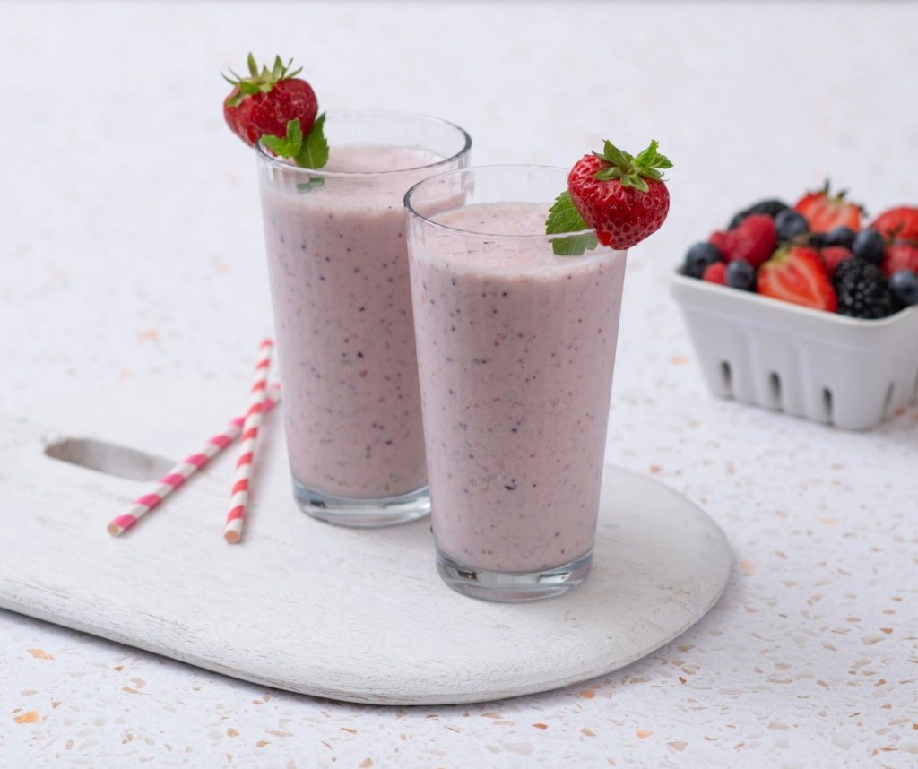 Two glasses of pink smoothies with strawberries and mint garnish, on a wooden board, next to a basket of mixed berries.