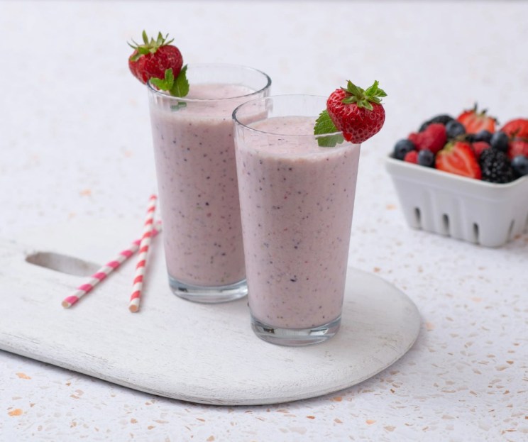 Two glasses of pink smoothies with strawberries and mint garnish, on a wooden board, next to a basket of mixed berries.