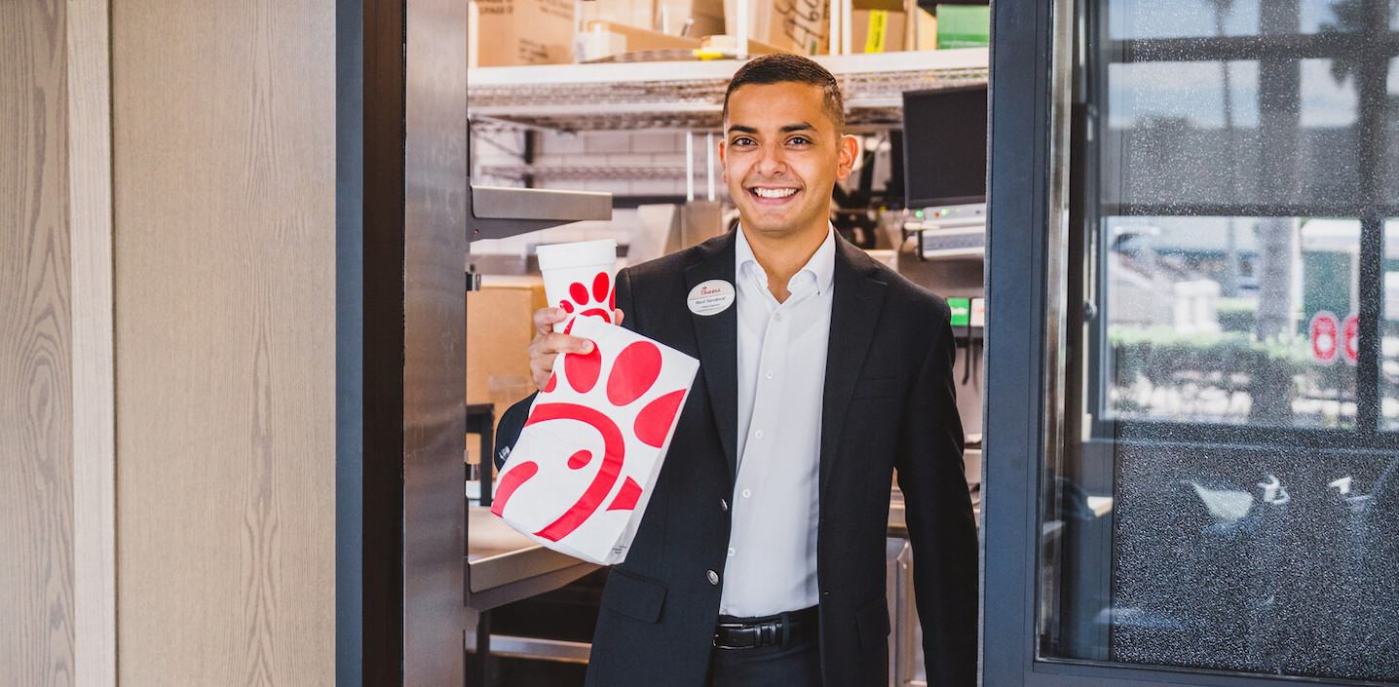 A person in a black suit holding a Chick-fil-A branded cup and bag inside a Chick-fil-A restaurant.