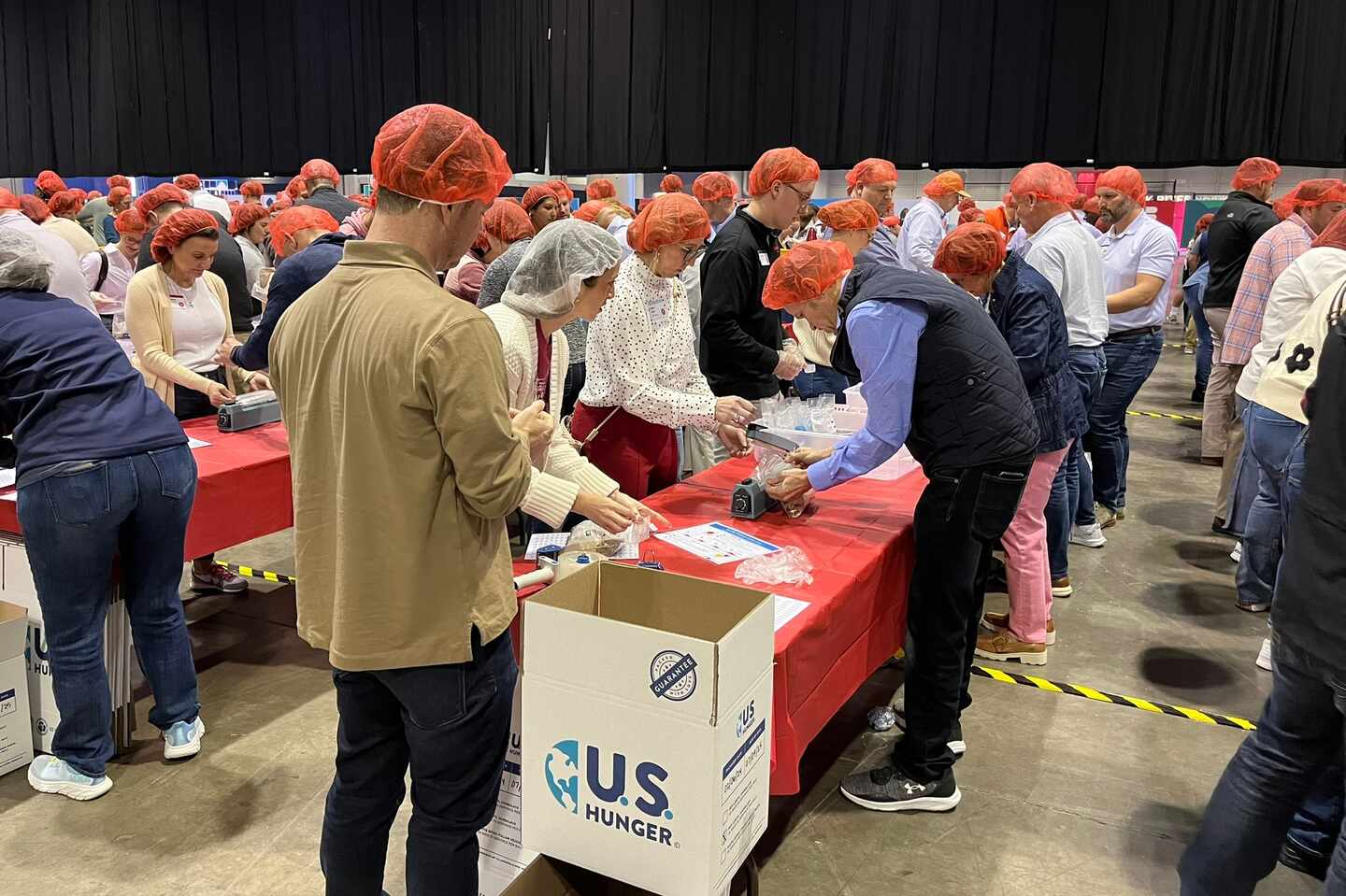 People wearing red hairnets participate in a meal-packing event at long red-covered tables, with a U.S. Hunger box in the foreground.