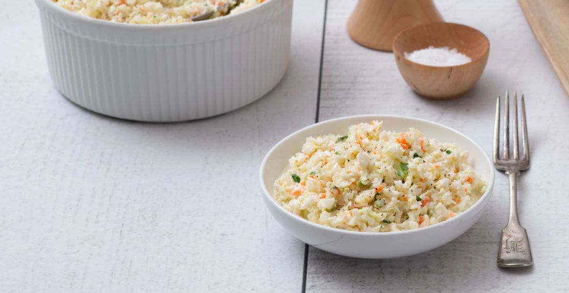 A serving bowl of cole slaw with an individual serving plated next to it with a fork