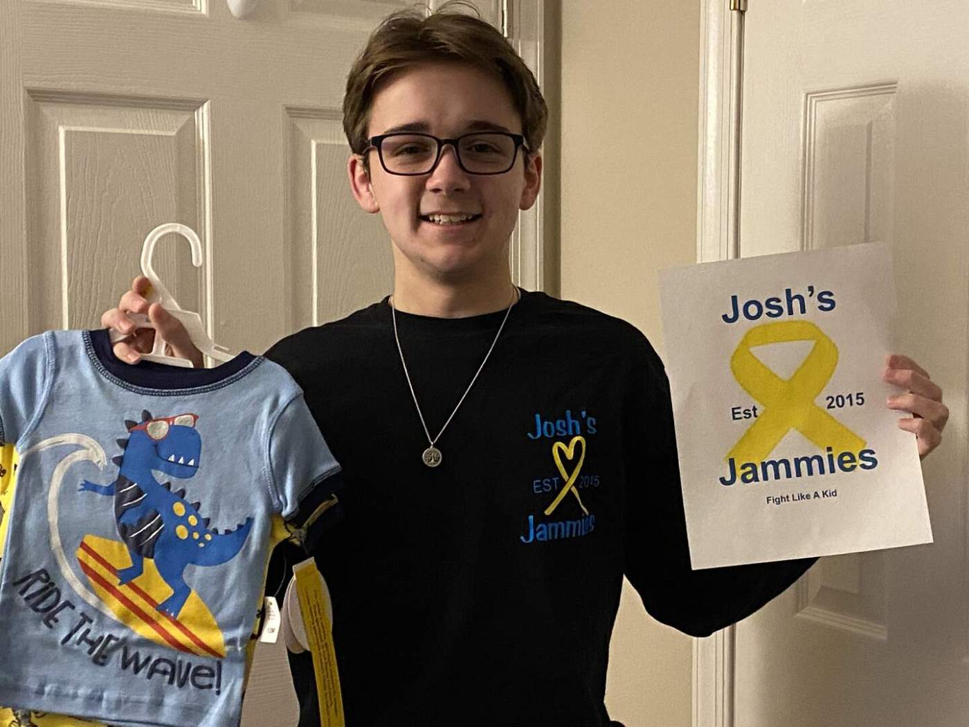 Person holding a dinosaur-themed pajama on a hanger and a paper with yellow ribbon design, with the text Josh’s Est 2015 Jammies Fight Like A Kid.