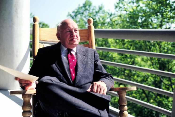 S. Truett Cathy sitting in a rocking chair outside and holding a paper.