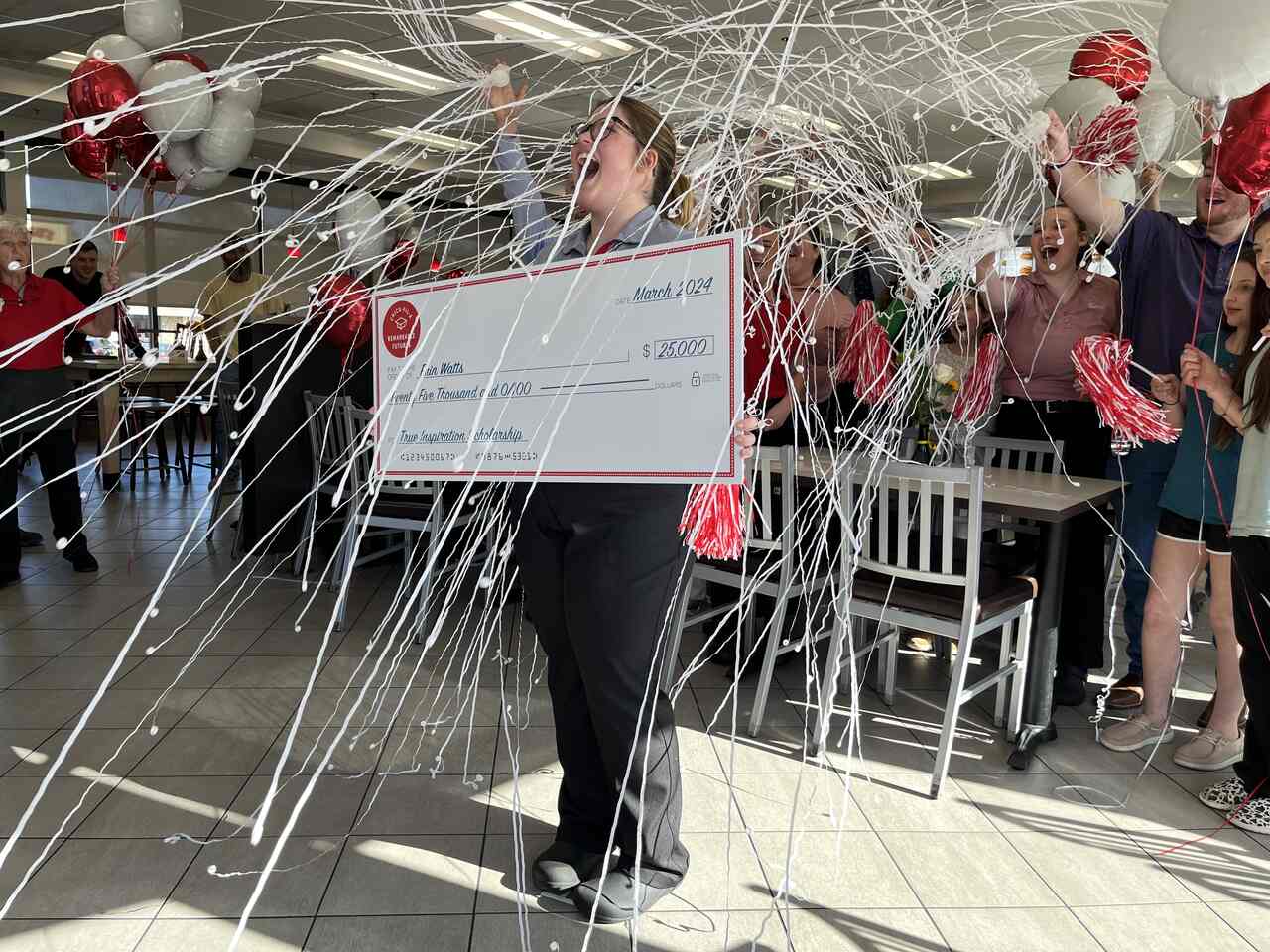 A team member holds an oversized check while her team members celebrate with balloons and streamers.