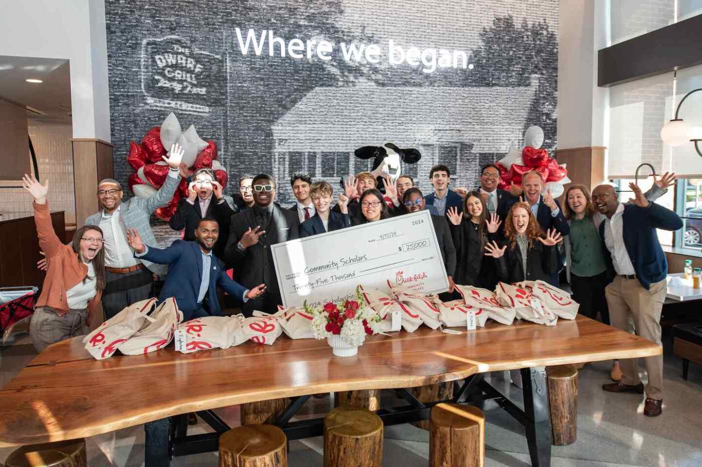 A group celebrating with an oversized check and gift bags in front of a mural reading "Where we began".