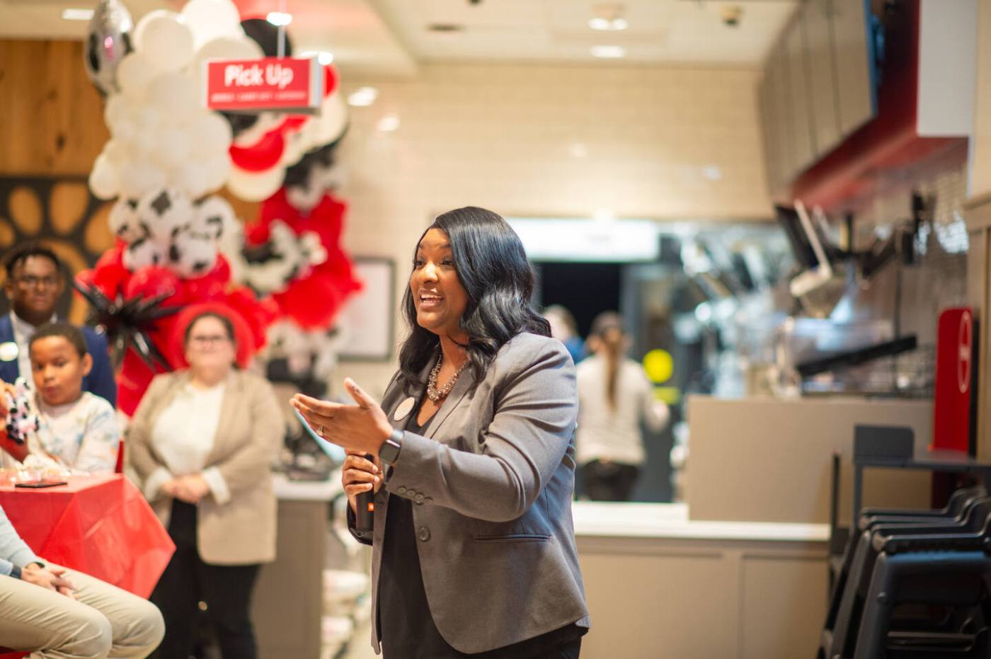 Katie Branch speaks to guests during a Chick-fil-A event, standing near red and white balloon decorations and a 'Pick Up' sign.