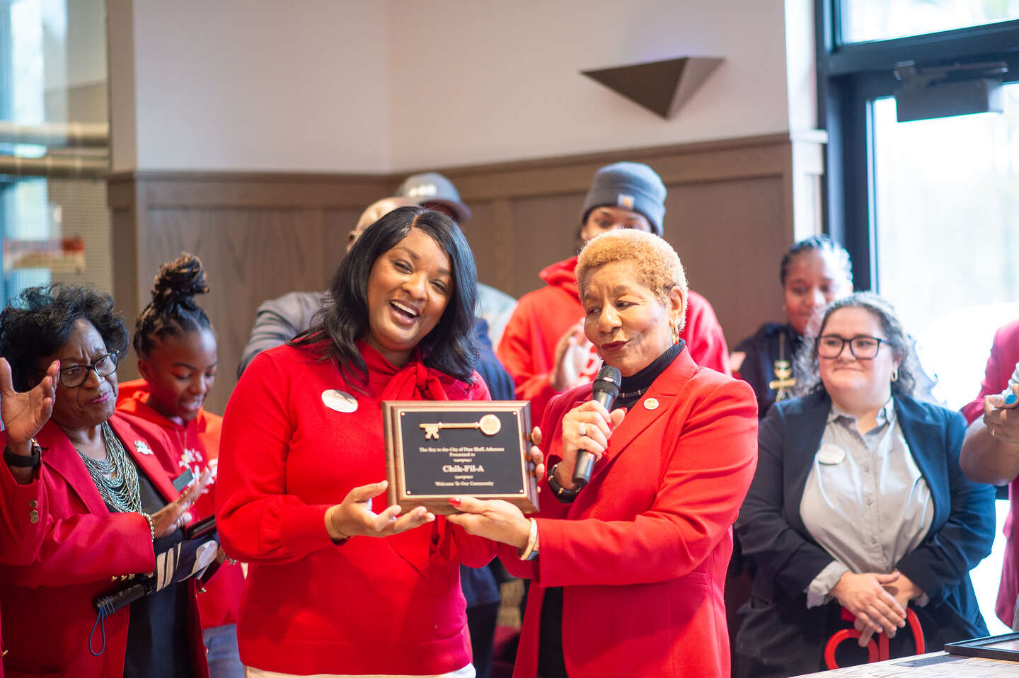 Katie Branch receives a plaque and smiles alongside a woman speaking into a microphone at a Chick-fil-A community celebration.