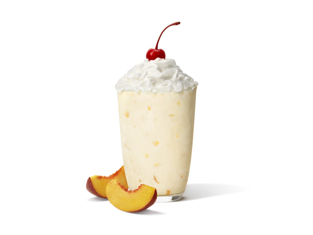 A peach milkshake in a glass with whipped cream and a cherry, next to fresh sliced peaches.