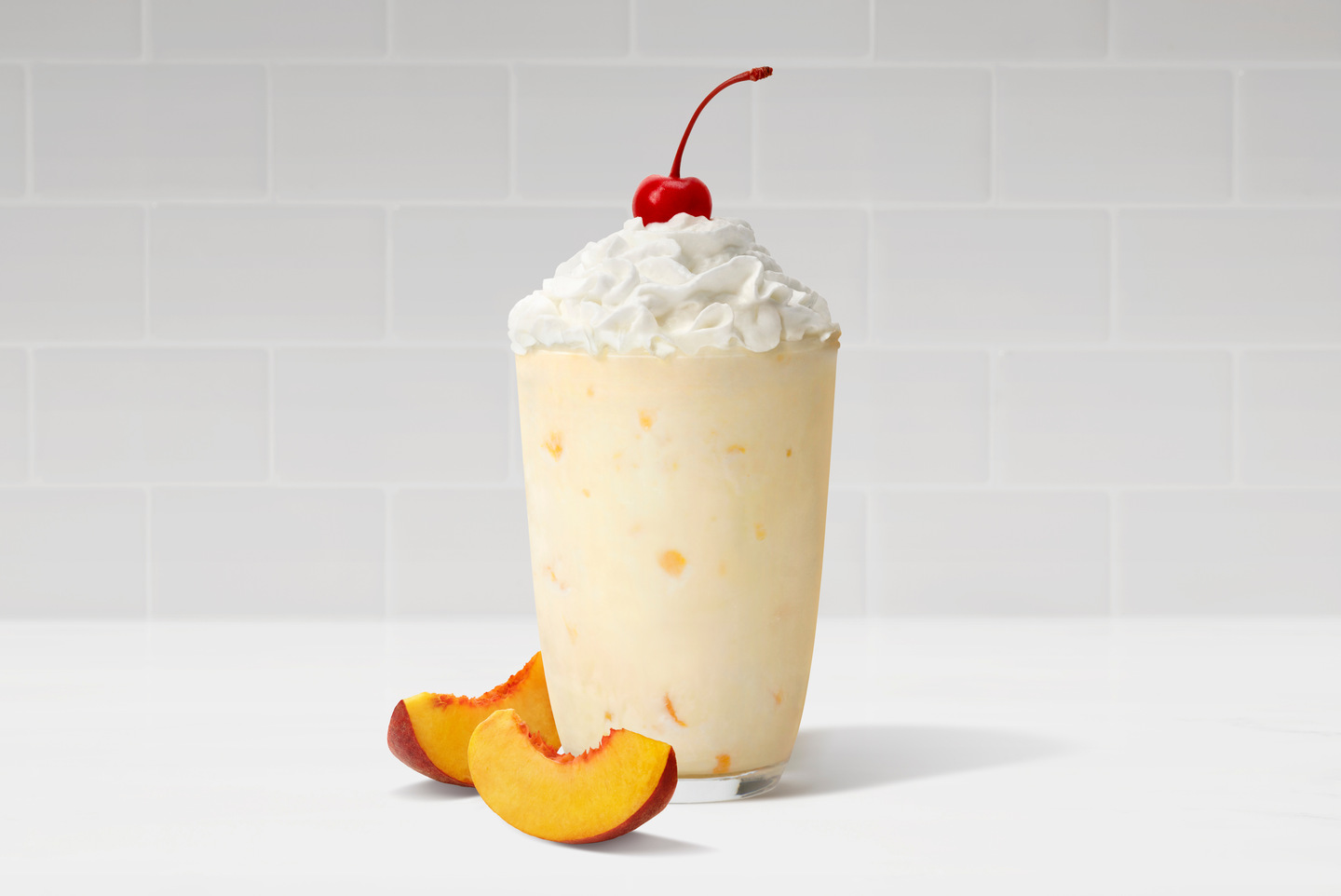 A peach milkshake in a glass with whipped cream and a cherry, next to fresh sliced peaches.
