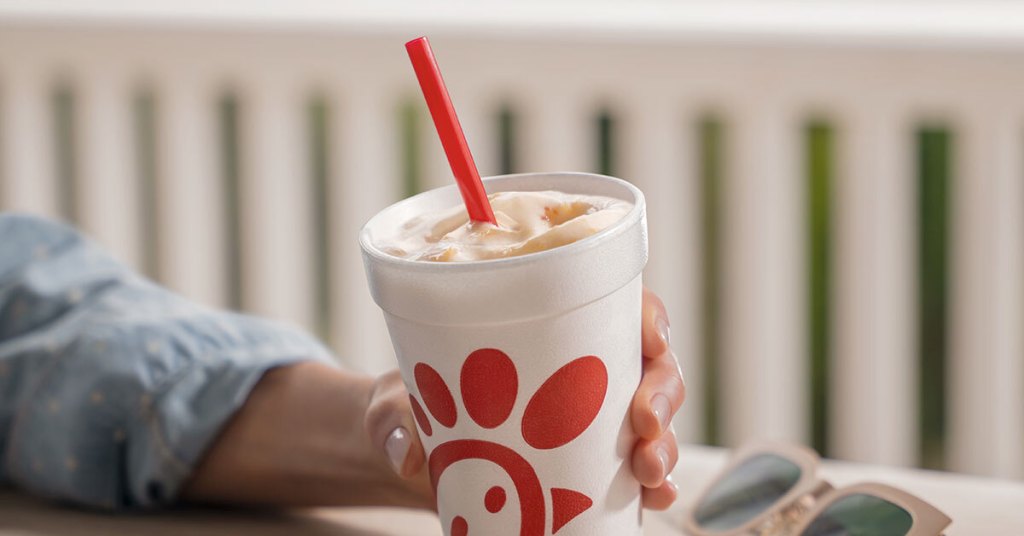 A hand holding a foam cup with a peach milkshake and a red straw.